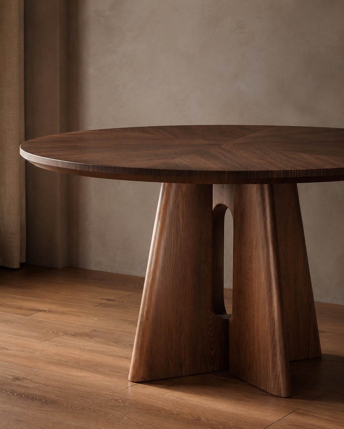 A round dark wood dining table with a sculptural pedestal base, set in a warm, minimalist room with a textured beige wall and wooden floor.