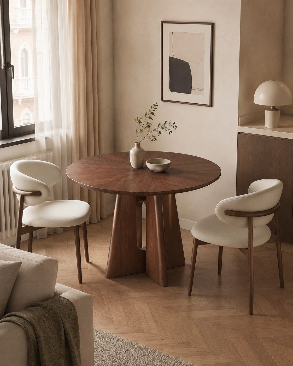 A round wooden dining table with a sculptural pedestal base, accompanied by two modern white upholstered chairs with curved wooden arms, set in a warm, minimalist dining room with beige walls and herringbone wood flooring.